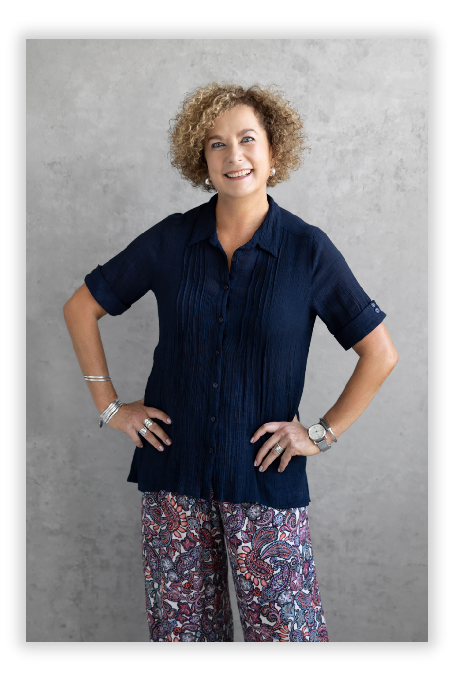 Sonja Cronje, a professional coach for healthcare professionals stands smiling against a gray background with curly hair. She’s wearing a navy blue blouse and patterned pants, accessorized with bracelets and a watch, with hands on her hips.